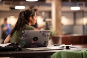 Laptop of security researcher covered customer stickers. Young woman watching intently. 